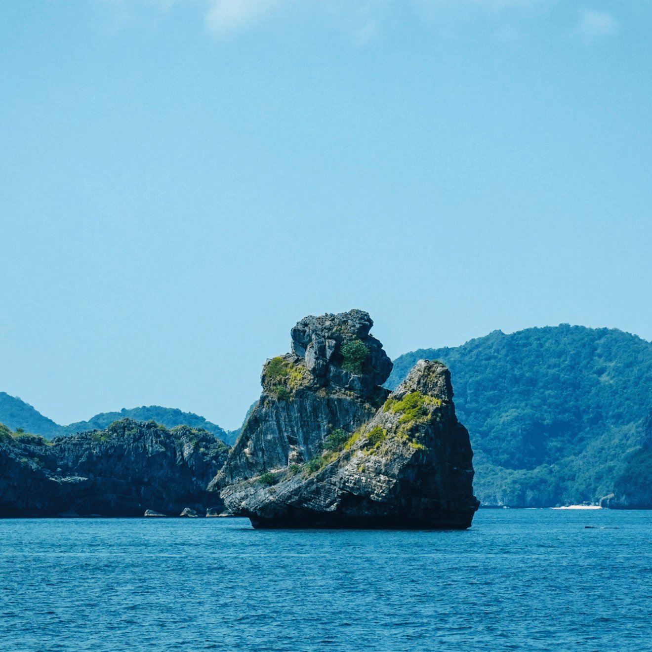 Rocky island in blue sea with distant green hills under clear sky.
