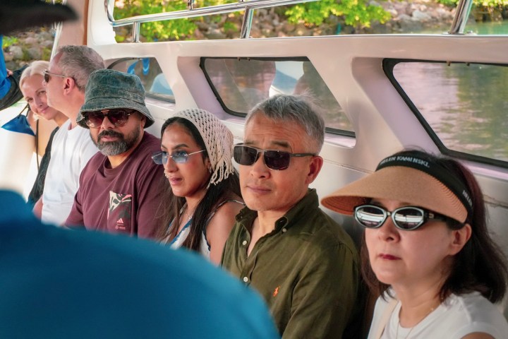 Passengers seated inside a boat, wearing hats and sunglasses, with trees visible outside.