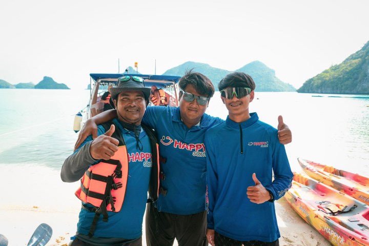 Three people smiling on a beach near kayaks, wearing blue shirts and sunglasses.
