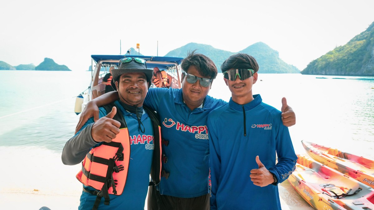 Three people smiling on a beach near kayaks, wearing blue shirts and sunglasses.