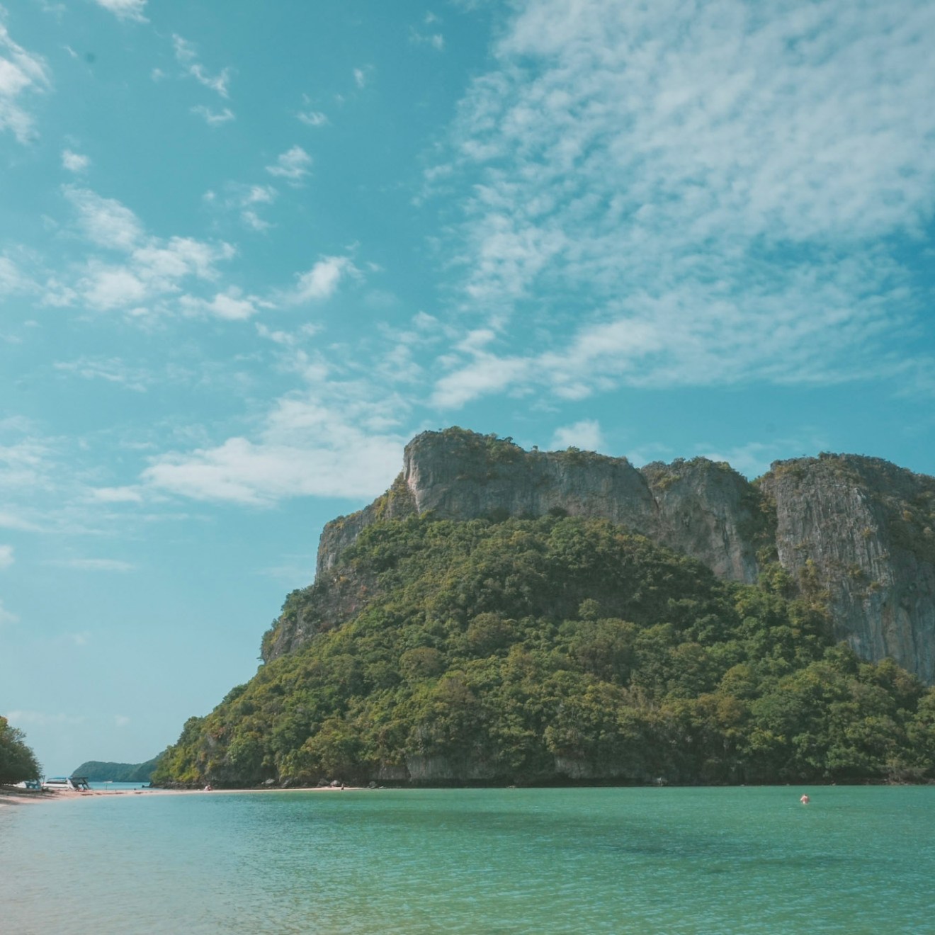 Serene beach with clear water and a large, lush green rocky hill under a blue sky with clouds.
