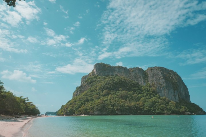 Serene beach with clear water and a large, lush green rocky hill under a blue sky with clouds.