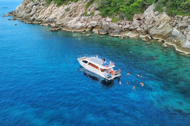 Boat anchored near rocky coastline with people swimming in clear blue water.