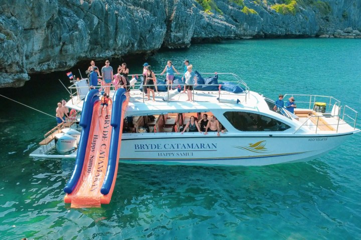 A boat with a water slide and people onboard near rocky cliffs in clear water.