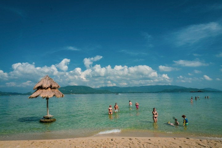 Beach scene with people swimming, straw hut, and mountains in the background.