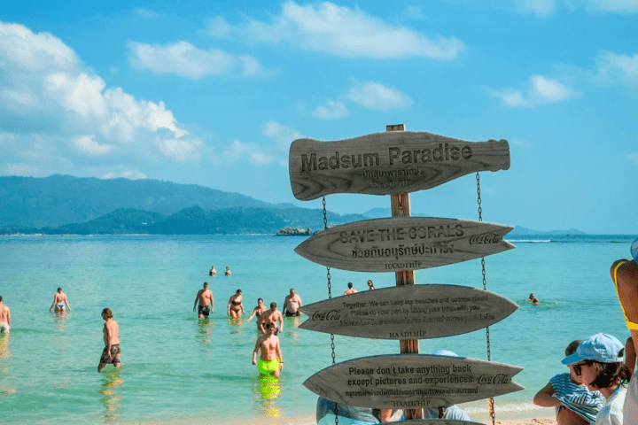 Beach scene with people swimming and a wooden signpost on the sand under a blue sky.