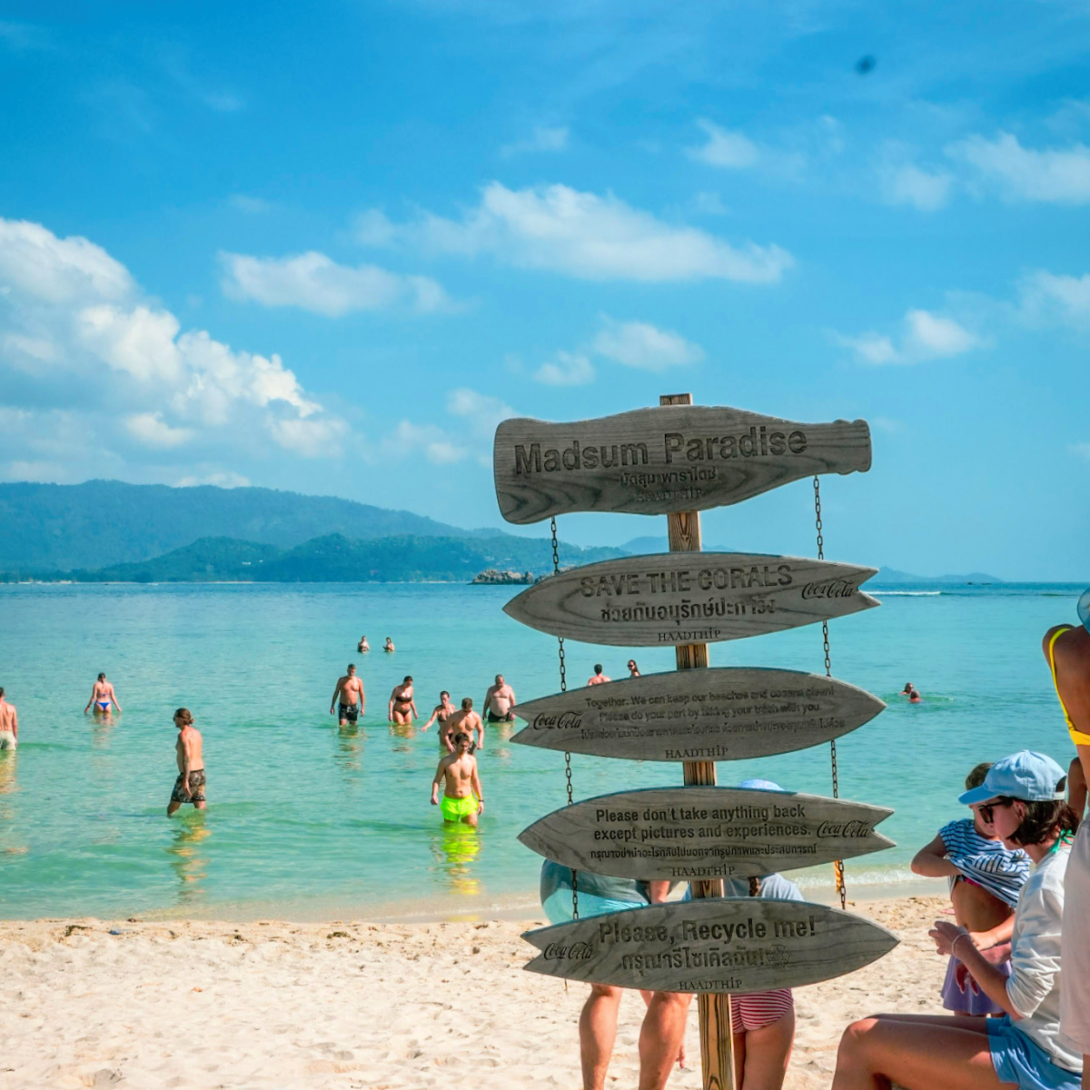 Beach scene with people swimming and a wooden signpost on the sand under a blue sky.