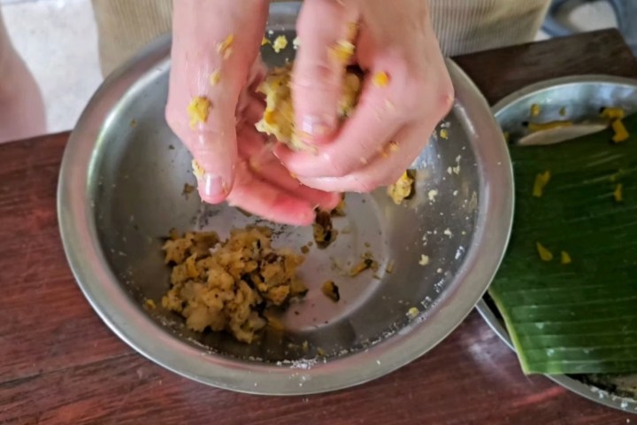 Hands mixing dough in a metal bowl next to a banana leaf.