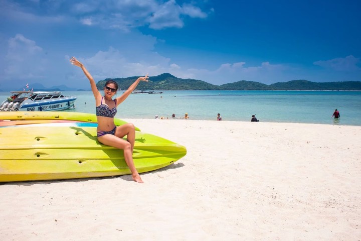 Woman in swimsuit sitting on yellow kayak on a sunny beach, with ocean and mountains in background.