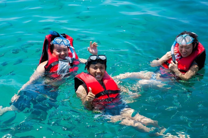 Three people in life vests and snorkels float in clear blue water, smiling and waving.