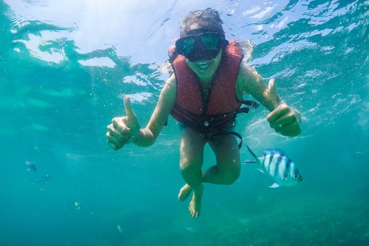 Person snorkeling underwater, smiling with thumbs up, wearing life vest and goggles, with fish swimming nearby.