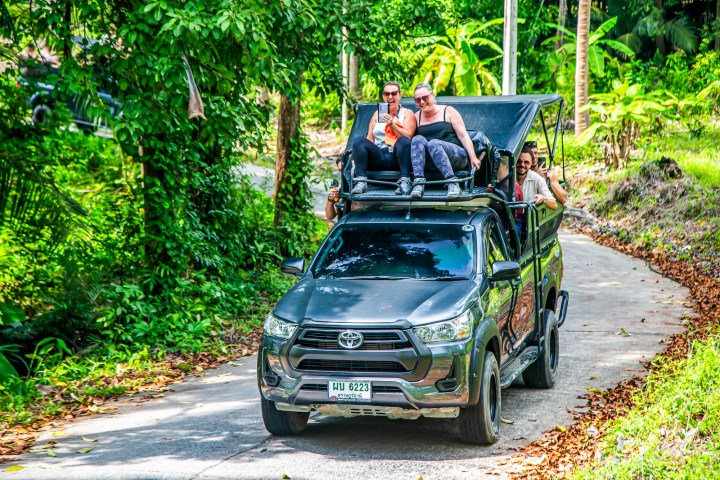 People riding on roof of SUV on a lush rural road.