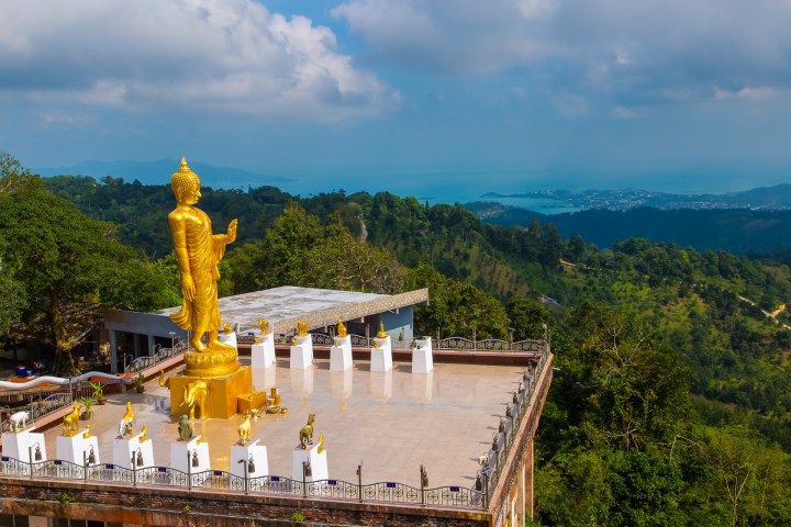 Golden Buddha statue overlooking a lush, green landscape with mountains and a cloudy sky.