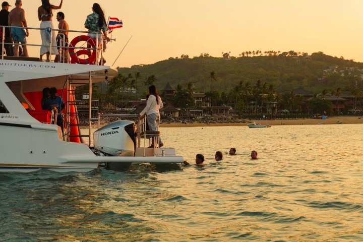 People on a boat and swimming in water near a tropical beach at sunset.
