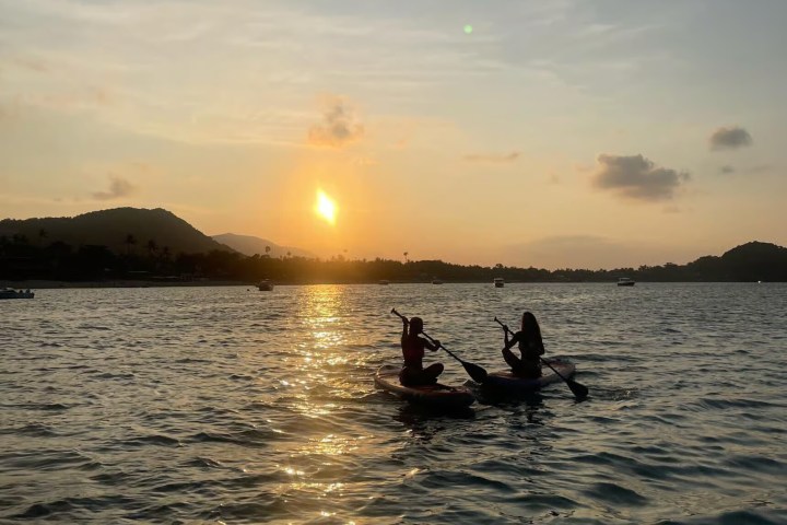 Two people kayaking on a calm sea at sunset with mountains in the background.