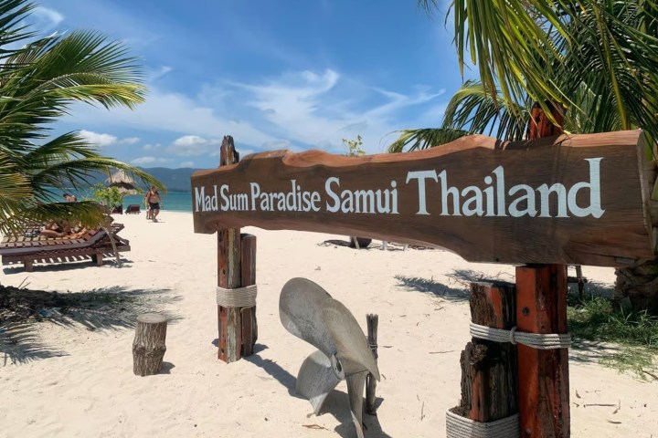 Beach with sand, palm trees, and sign reading 'Mad Sum Paradise Samui Thailand'.
