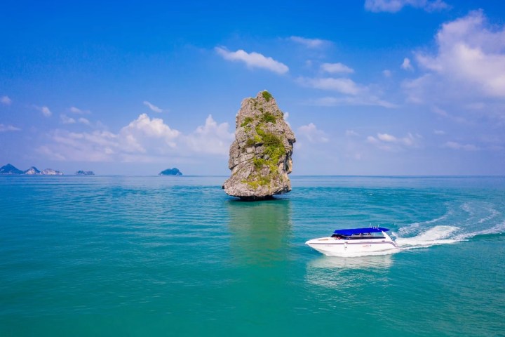 Speedboat on blue sea near tall rock formation under a sunny sky.