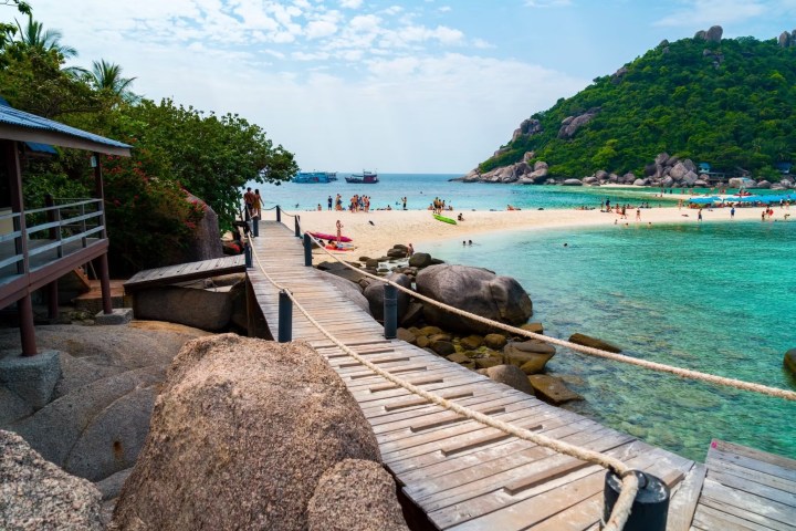 Wooden walkway leads to tropical beach with people, clear water, and a lush island.