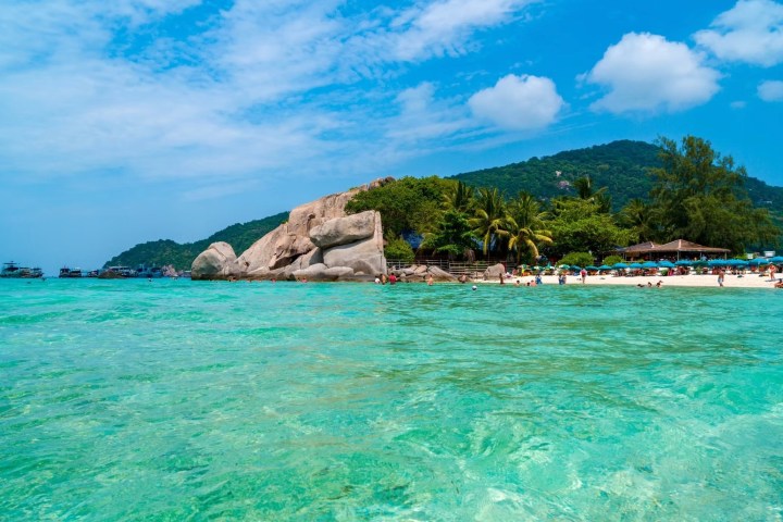 Clear turquoise water, rocky shoreline, and lush greenery with blue sky.