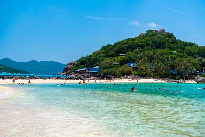 Scenic beach with turquoise water, people swimming, green hills, and clear blue sky.