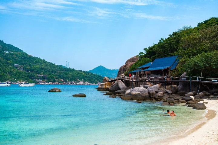Tropical beach with clear water, rocks, and a wooden structure beside lush green hills under a blue sky.