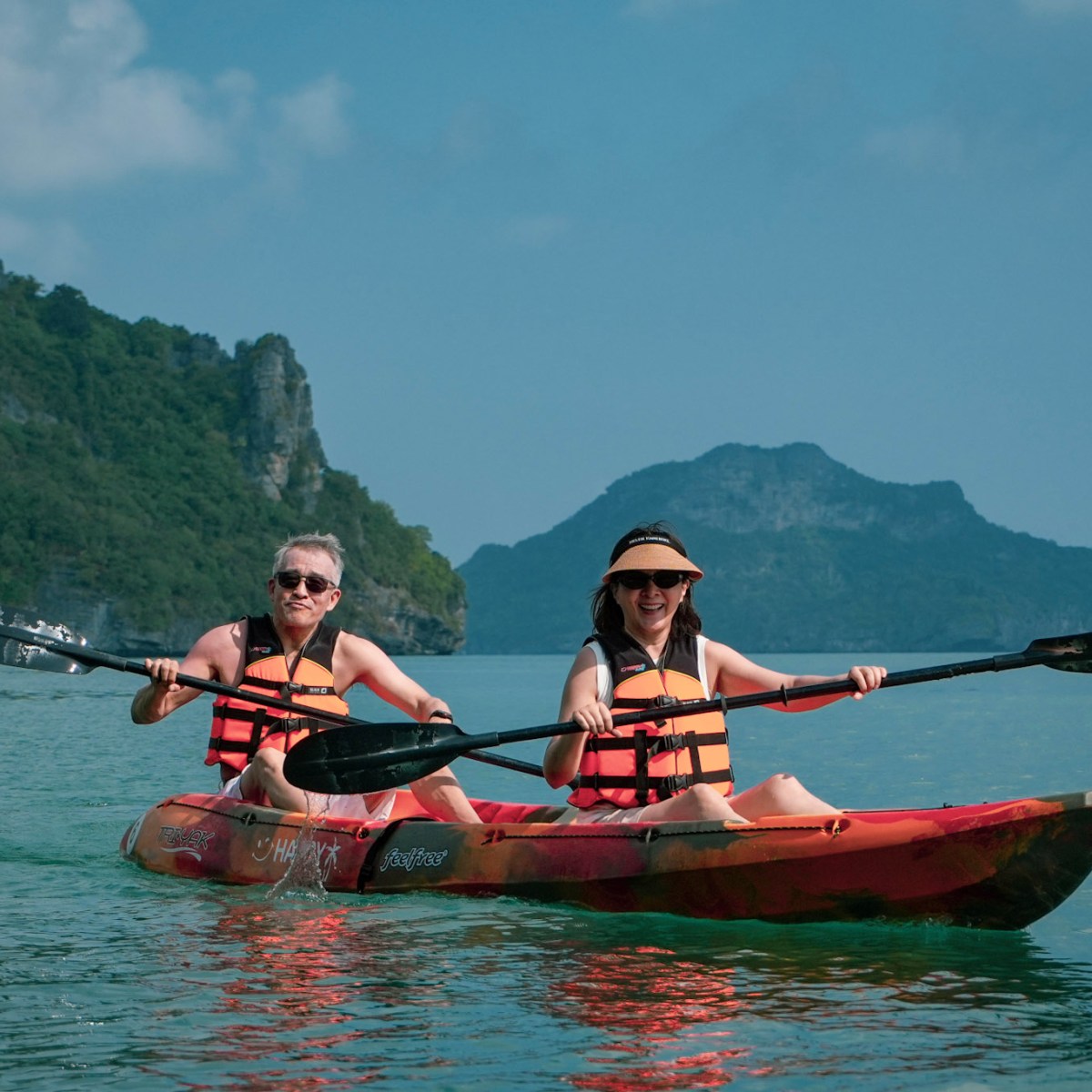 Two people in life vests kayaking on calm water near rocky islands.
