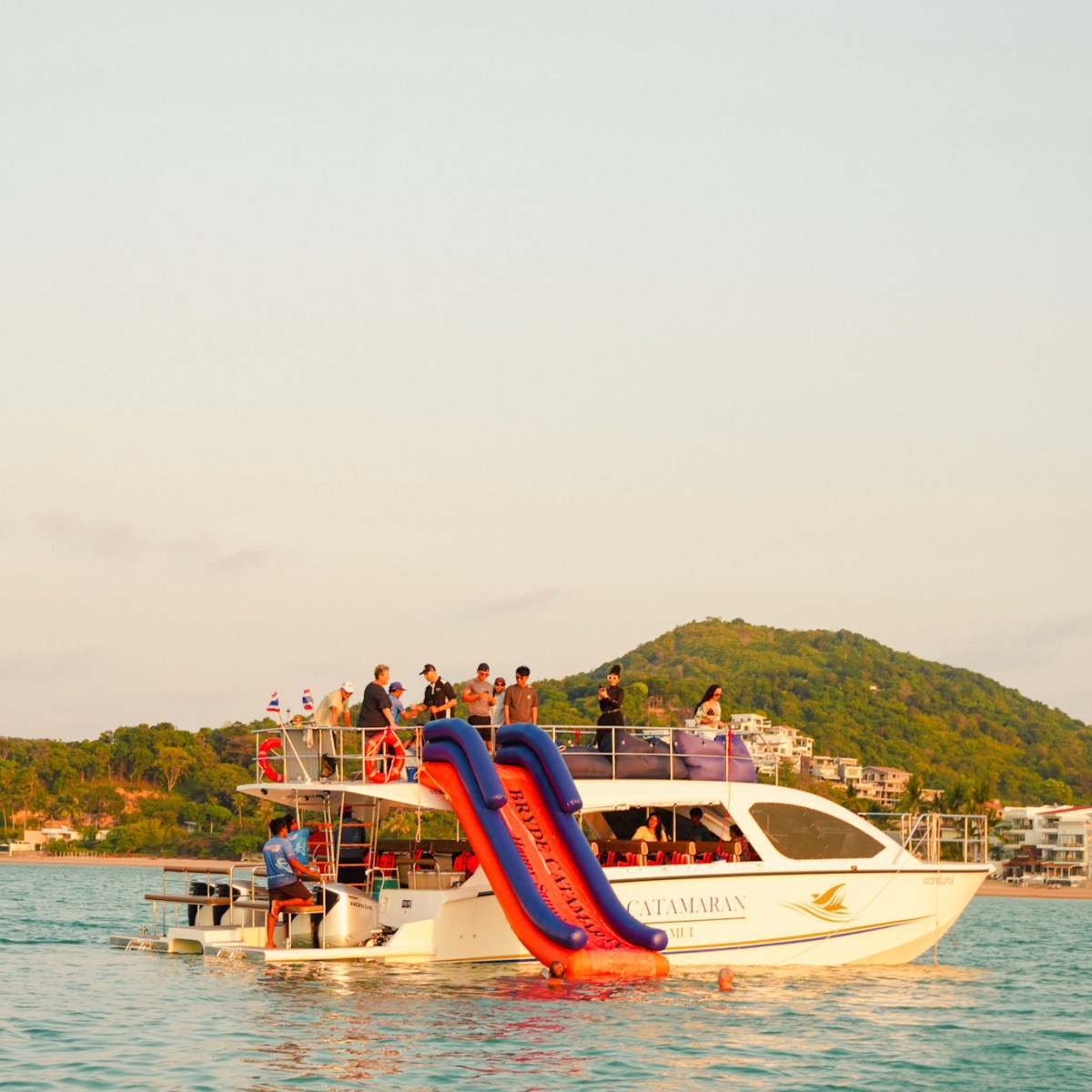 Boat with a slide in a bay, people onboard, hill and buildings in background.
