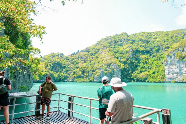 People on a viewing platform overlooking a green lagoon and forested hills.