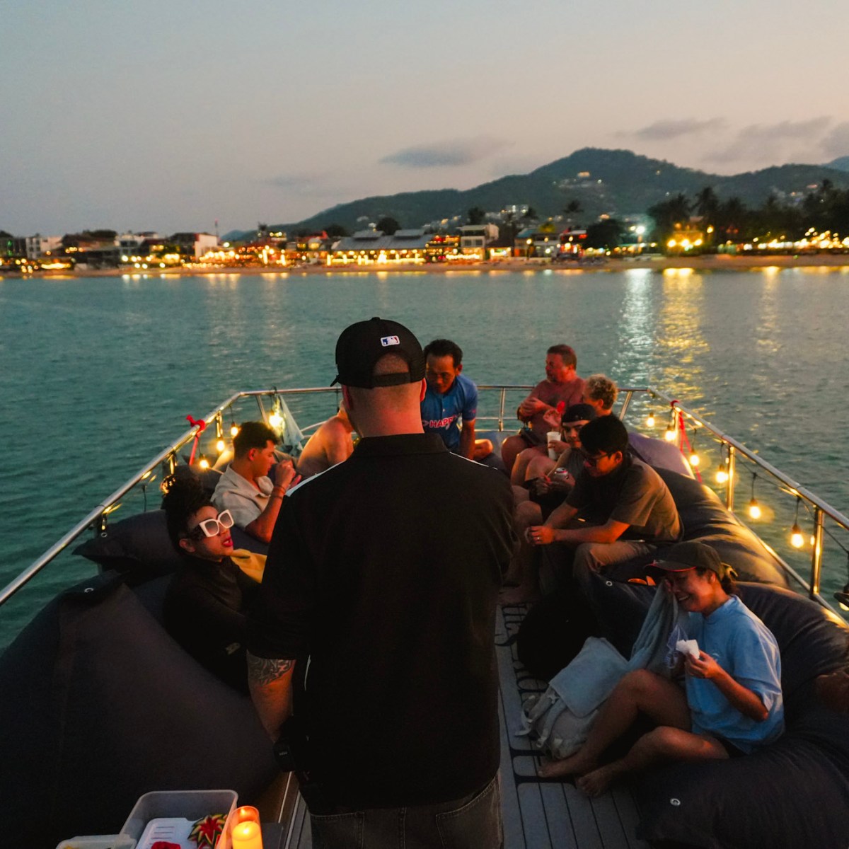 Group of people enjoying a boat ride at dusk, with lights and scenic coastline in the background.