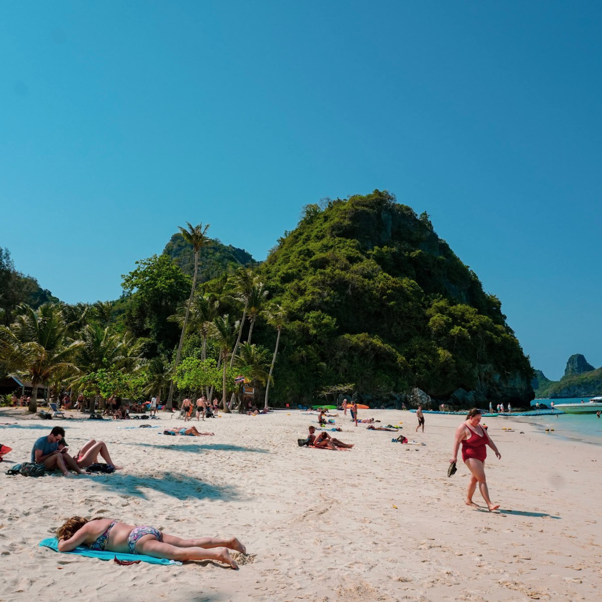 People relax on a sandy beach with lush green hills and clear blue sky.