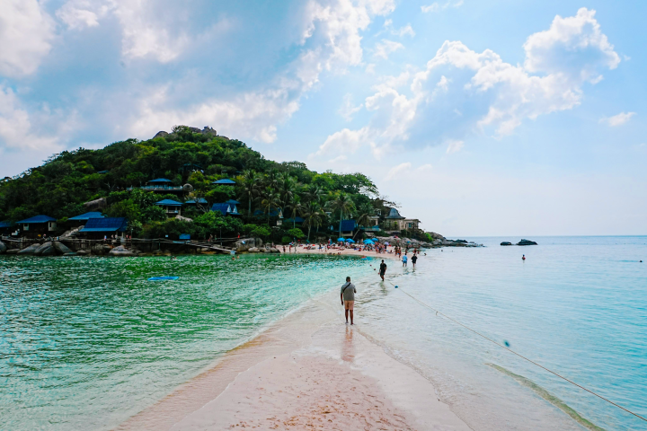 People walk on a sandbar towards a lush island with buildings under a cloudy sky.