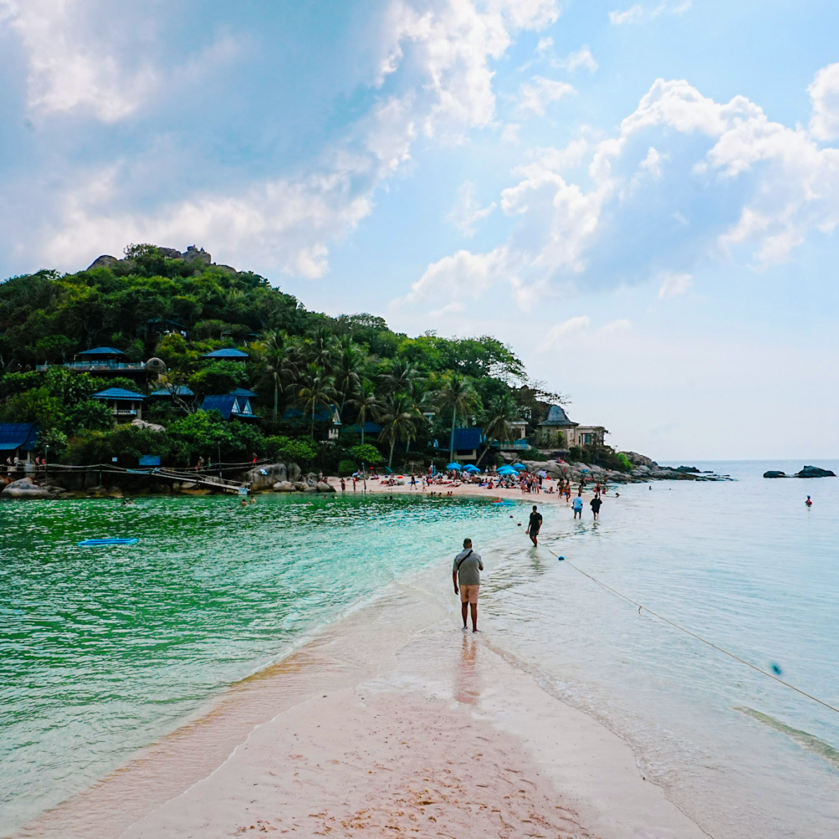 People walk on a sandbar towards a lush island with buildings under a cloudy sky.