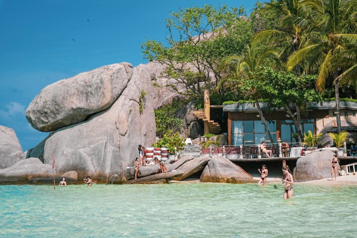 Beach with rocks, palm trees, people swimming, and building near the shore.