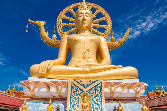 Golden Buddha statue under blue sky, with a person kneeling in prayer.