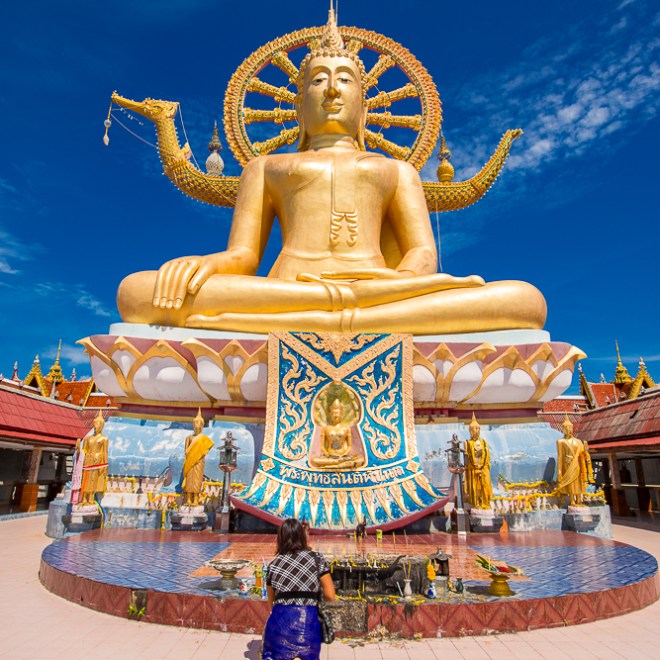 Golden Buddha statue with intricate details under a blue sky, person kneeling in prayer at base.